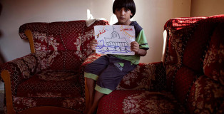 Il futuro nei disegni dei bambini di Gaza. 6-year old Yosef Al Yazir, seated on the arm of a chair in the living room of his home in Gaza City’s Ramal neighbourhood, holds a drawing showing his view of Gaza in the future. He remembers the last war, but states that this one is much worse: “There are a lot of children killed,” he said. (Image credits: Aya © UNICEF/NYHQ2014-1443/d’Aki)