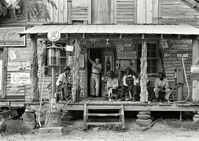 Dorothea Lange - Negozio di campagna su strada sterrata. Domenica pomeriggio. Si noti la pompa del cherosene a destra e quella della benzina a sinistra. I pali di legno grezzi e non rifiniti sono stati usati come sostegno per il tetto della veranda. Uomini neri sono seduti sulla veranda. Il fratello del proprietario del negozio è in piedi sull’ingresso Gordonton, North Carolina. 1939 The New York Public Library | Library of Congress Prints and Photographs Division Washington.