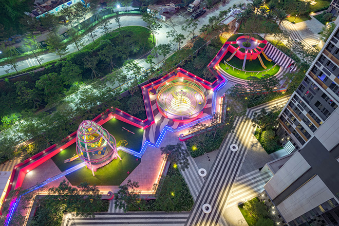 Ballistic Architecture Machine (BAM) - Buji Residences. Aerial dusk view of the multiple playgrounds at Buji Residences connected by a meandering terrain wall in bold pink and purple and a central blue LED river element connecting waterplay features. Photo credit: Amey Kandalgaonkar
