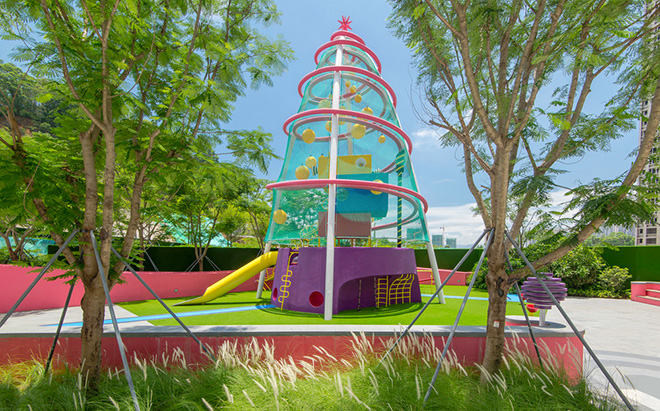 Ballistic Architecture Machine (BAM) - Buji Residences. Playscraper playground at Buji Residences, with purple base and pink rings enclosing a blue mesh frame studded with yellow climbing modules and slide, set amidst lush planting beneath the towers. Photo credit: Amey Kandalgaonkar
